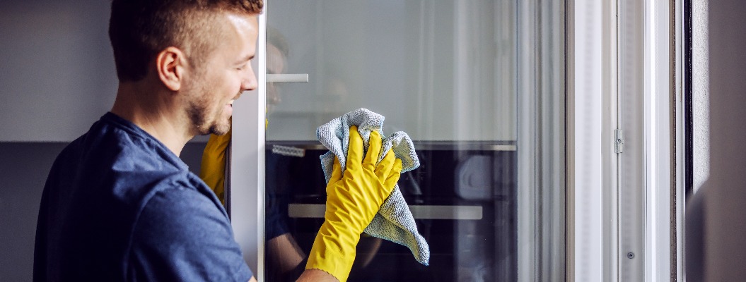 Junger bärtiger lächelnder positiver Mann mit Gummihandschuhen, der ein Fenster mit einem Tuch putzt. Nach Regen gibt es immer Flecken auf dem Glas.